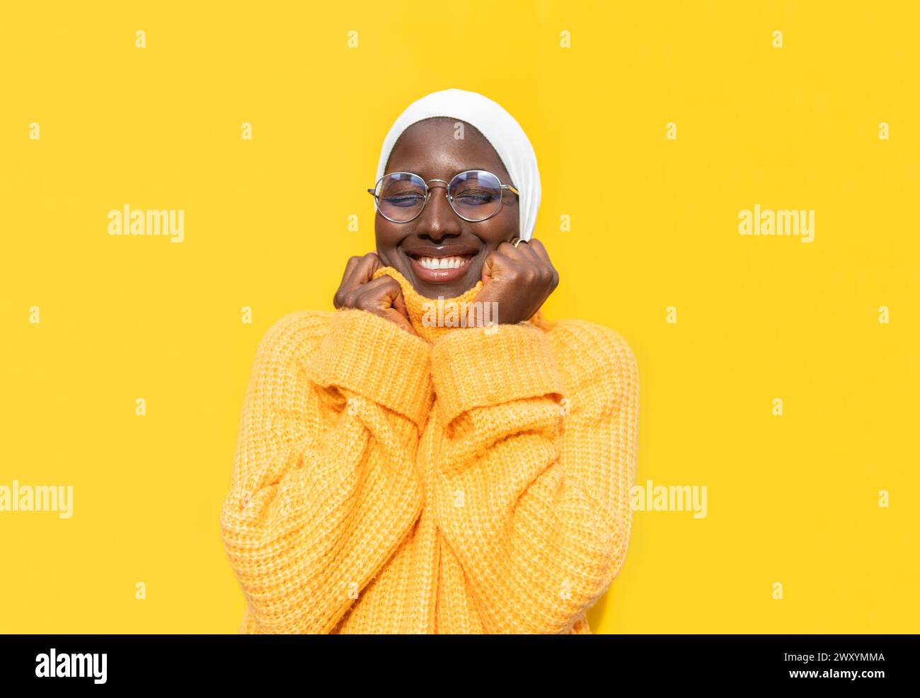 Una gioiosa donna afroamericana con gli occhi chiusi che indossa un hijab bianco e un comodo maglione giallo su uno sfondo giallo brillante, che trasuda felicità Foto Stock