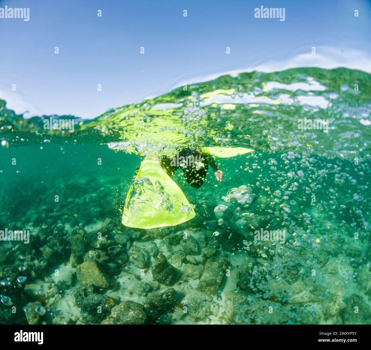 Una prospettiva subacquea dinamica di pinne gialle anonime dello snorkeling in mezzo a una raffica di bolle d'aria su un fondo di ciottoli in acque illuminate dal sole Foto Stock