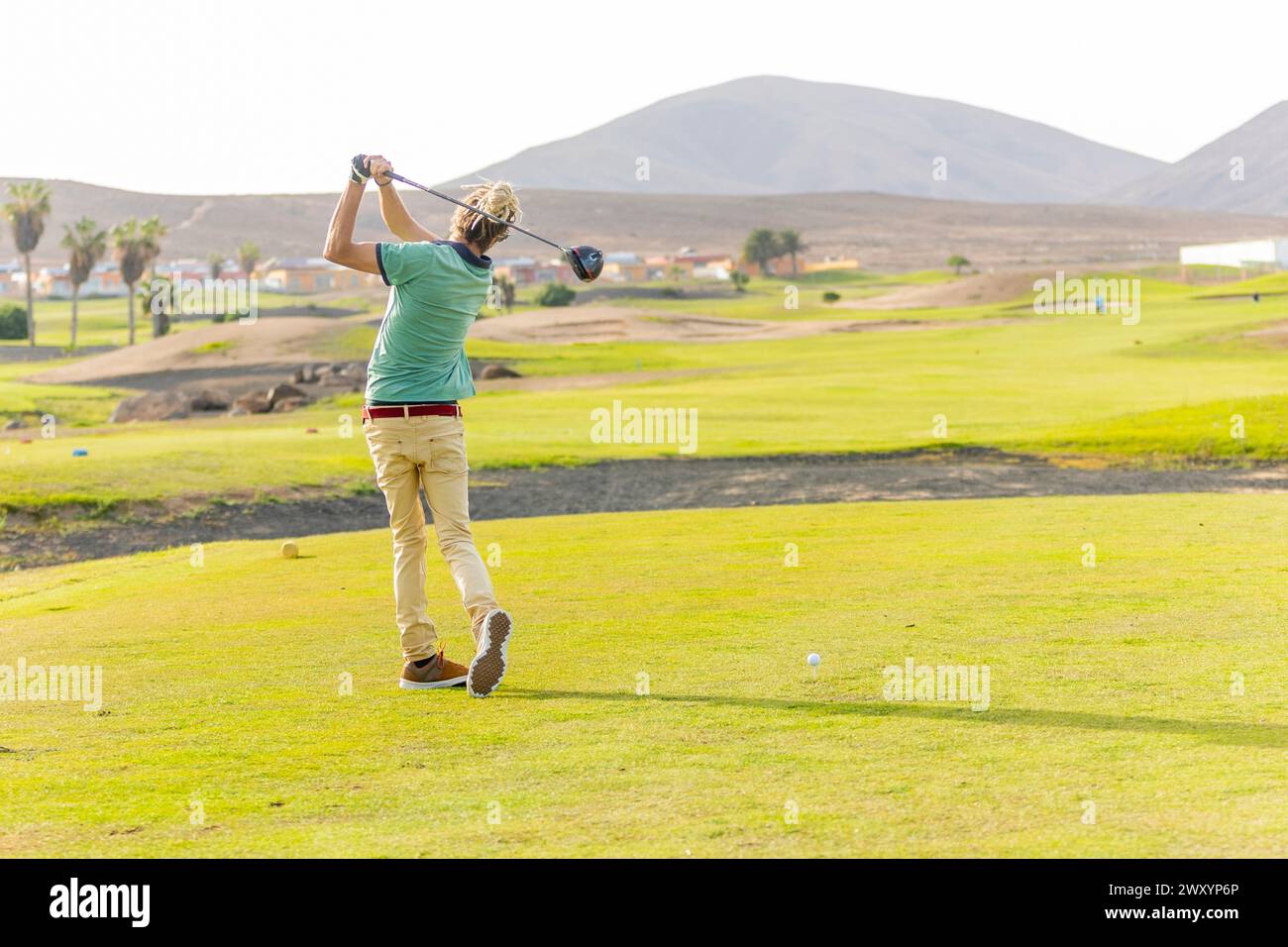 La vista posteriore di un golfista con dreadlock biondi viene catturata a metà corsa su un campo da golf panoramico, mostrando un aspetto non tradizionale nella spiaggia Foto Stock