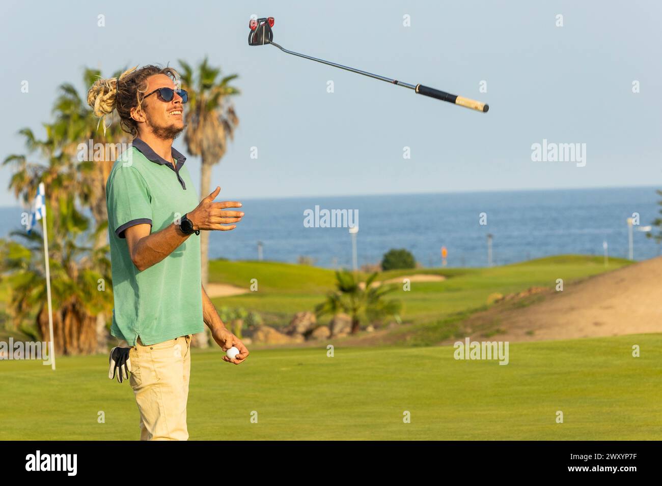 Un giovane golfista con i dreadlock fa scivolare casualmente la sua mazza dopo un'altalena su un pittoresco campo da golf sul mare. Foto Stock