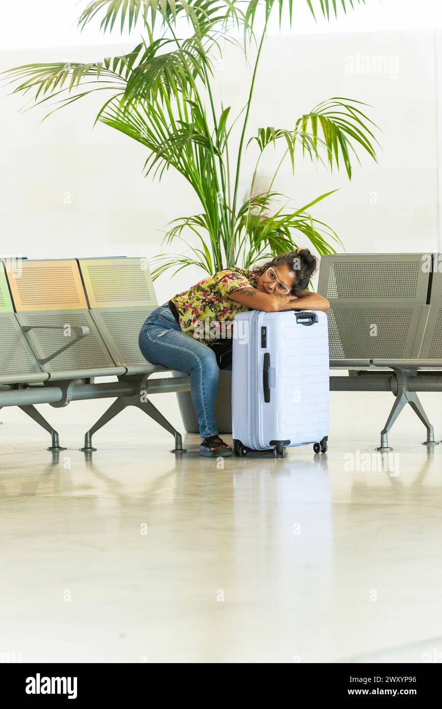 Un passeggero stanco si appoggia su una valigia bianca mentre attende nella sala partenze di un aeroporto, con una pianta tropicale e posti a sedere sullo sfondo Foto Stock