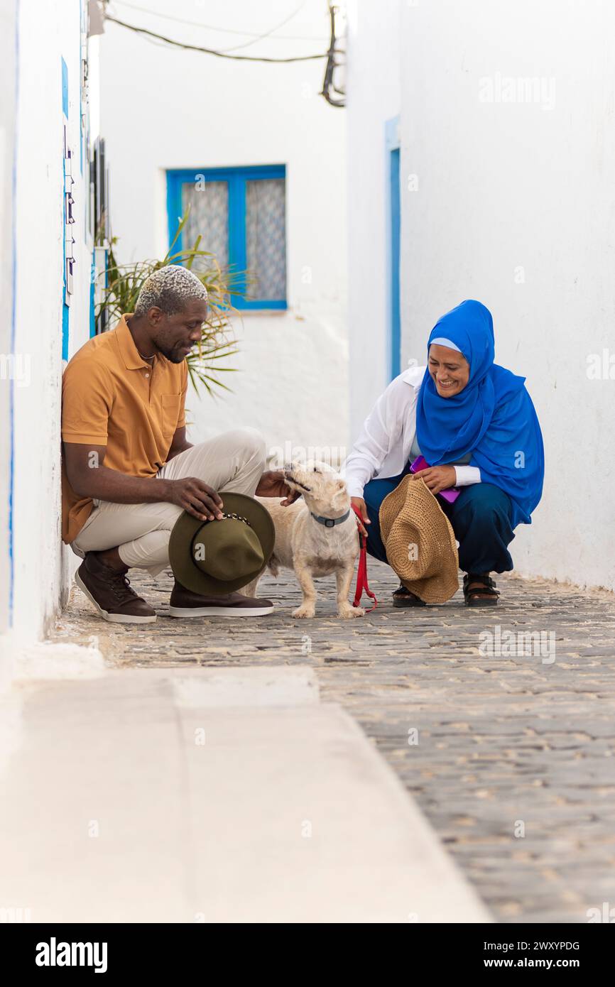 Una coppia multietnica sorride mentre accarezzano un cane allegro in una strada acciottolata, mostrando diversità e gioia Foto Stock