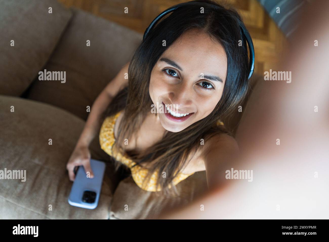 Una giovane donna sorridente che indossa le cuffie e tiene in mano uno smartphone scatta un selfie mentre si siede sul comodo divano di casa Foto Stock