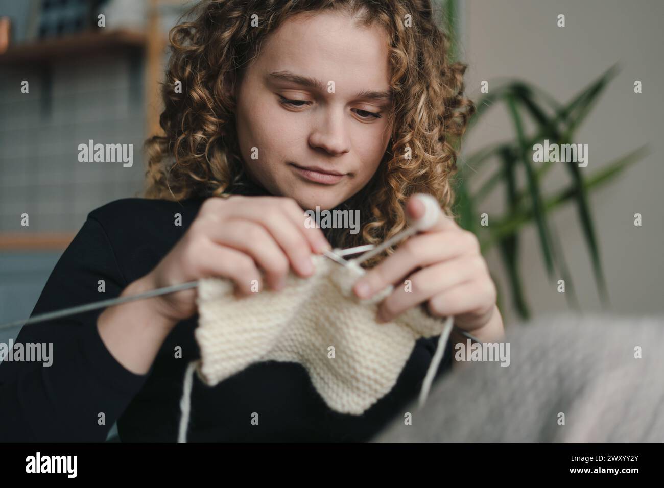 Giovane donna dai capelli ricci che lavora a maglia a mano mentre si siede sul divano a casa e si gode un hobby accogliente. Vestiti fatti a mano. La ragazza entra per lei Foto Stock
