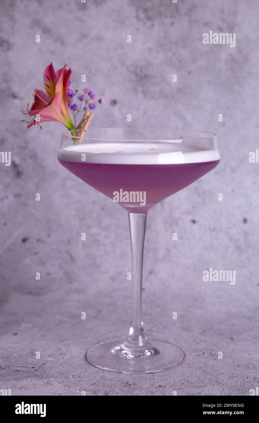 Elegante immagine di un cocktail rosa in un bicchiere con decorazione floreale su sfondo grigio, trasuda classe e raffinatezza. Foto Stock