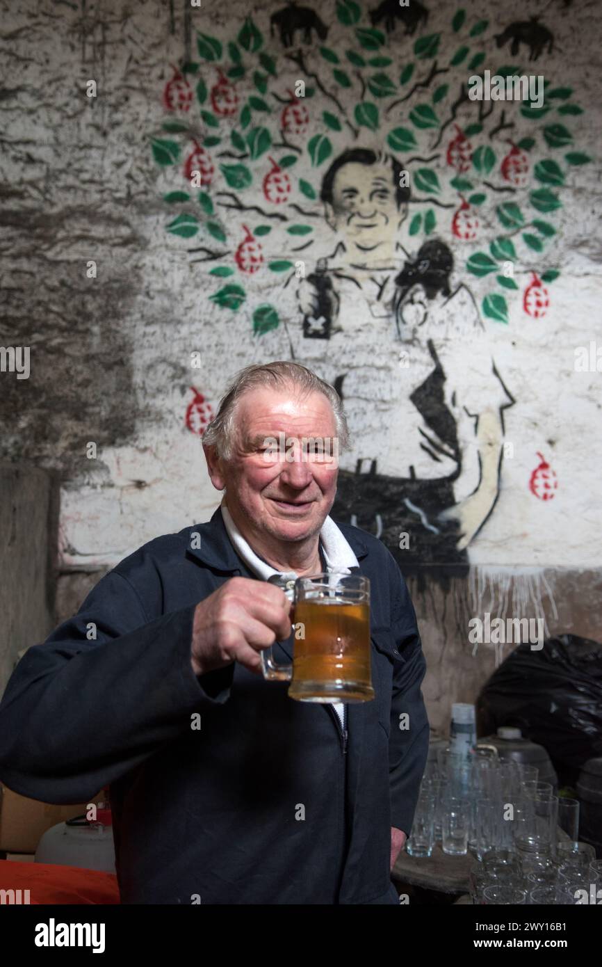 Roger Wilkins, un tradizionale coltivatore di sidro vecchio stile, in piedi di fronte a un ritratto Banksy che l'artista di graffiti ha fatto di lui. Roger beve una pinta di sidro secco. Ritratto situato in uno dei capannoni della fattoria. Land End Farm, Mudgley, Somerset, Regno Unito 2024 2020s HOMER SYKES Foto Stock
