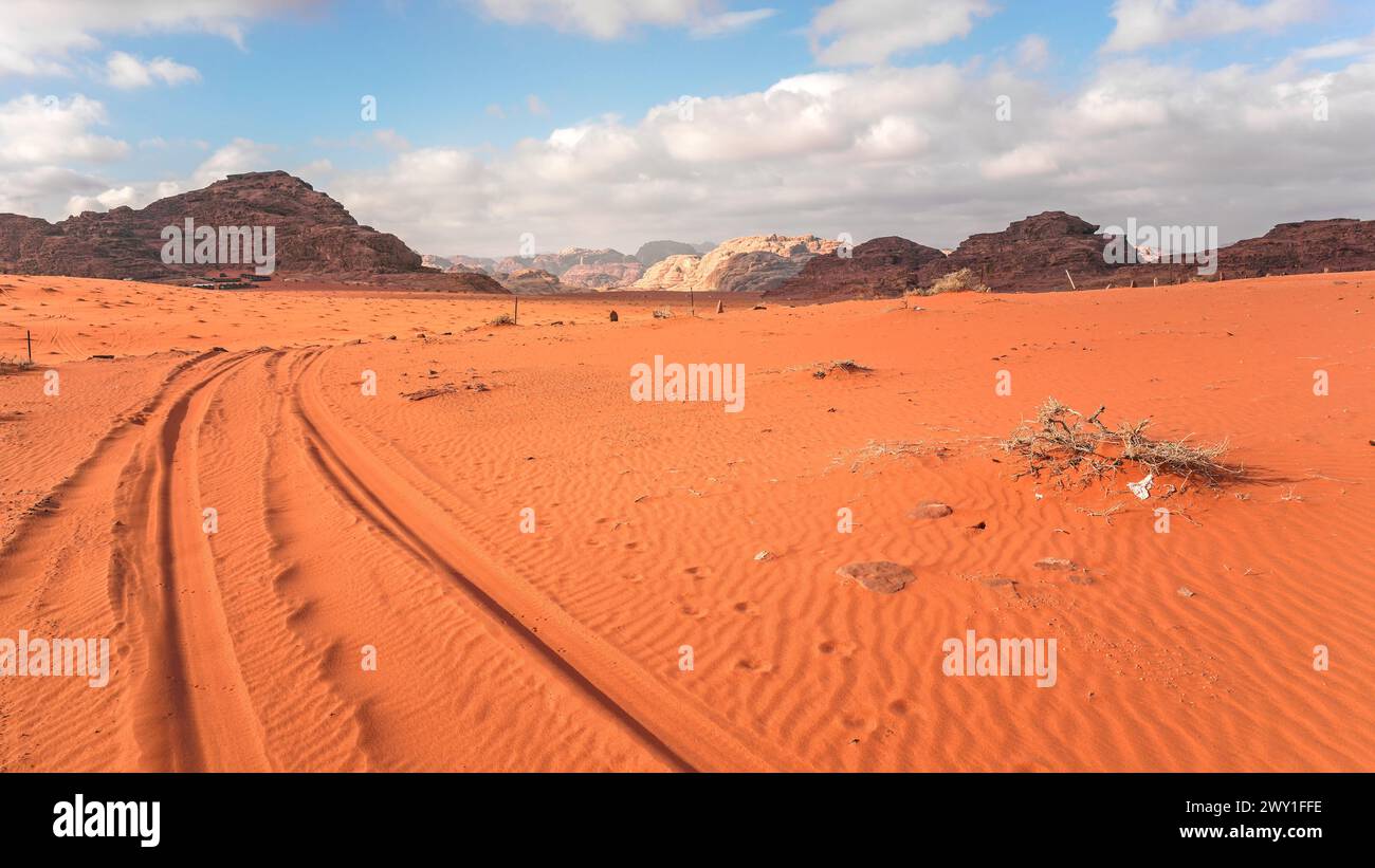 Massicci rocciosi sul deserto di sabbia rossa arancione, piste per veicoli, cielo nuvoloso luminoso sullo sfondo, tipico scenario di Wadi Rum, Giordania Foto Stock