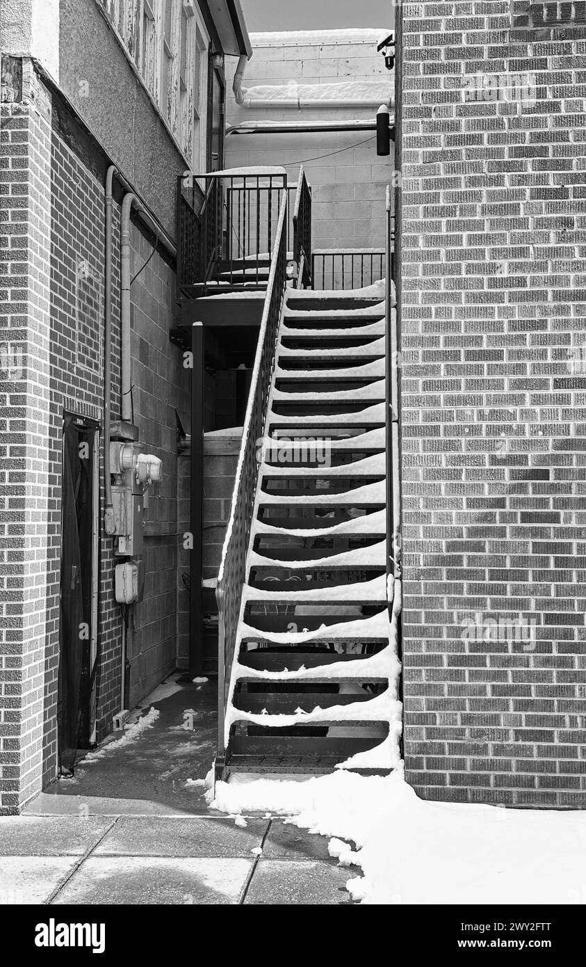 Il motivo bianco e nero monocromatico della neve sulle scale che salgono al secondo piano mostra il vicolo d'ingresso laterale alla zona commerciale in mattoni rossi della città. Foto Stock