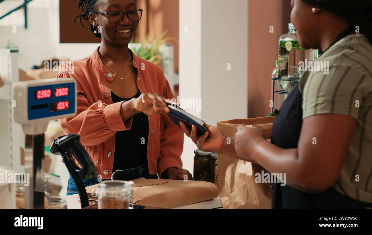 Cliente afroamericano che paga per prodotti biologici al momento del check-out, acquista prodotti di origine etica appena raccolti dal mercato agricolo locale. Acquirente che utilizza la carta presso il terminale POS, ecocompatibile. Foto Stock