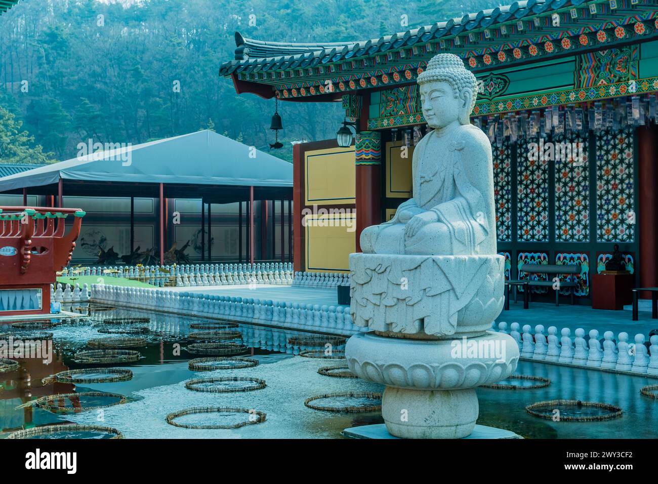 Grande Buddha seduto scolpito in pietra in un laghetto artificiale ghiacciato con l'edificio del tempio sullo sfondo a Yeongcheon, Corea del Sud Foto Stock