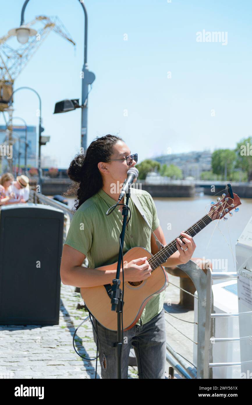 Immagine verticale di un giovane latino di etnia venezuelana, busker con capelli lunghi e occhiali, che lavora suonando la chitarra e cantando a Puerto Madero Arge Foto Stock