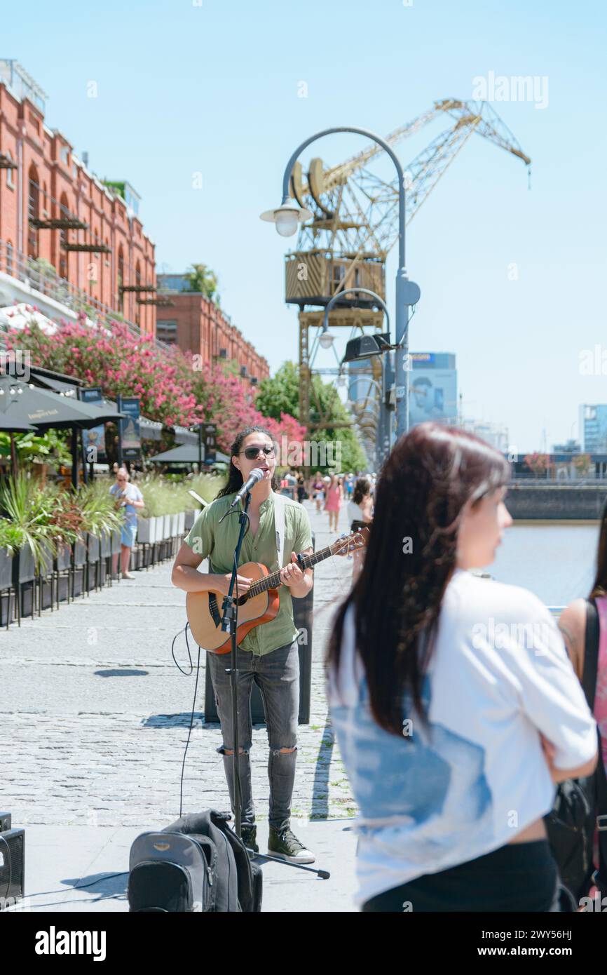 Immagine verticale di un giovane latino di etnia venezuelana, busker con capelli lunghi e occhiali, che lavora suonando la chitarra e cantando a Puerto Madero Arge Foto Stock