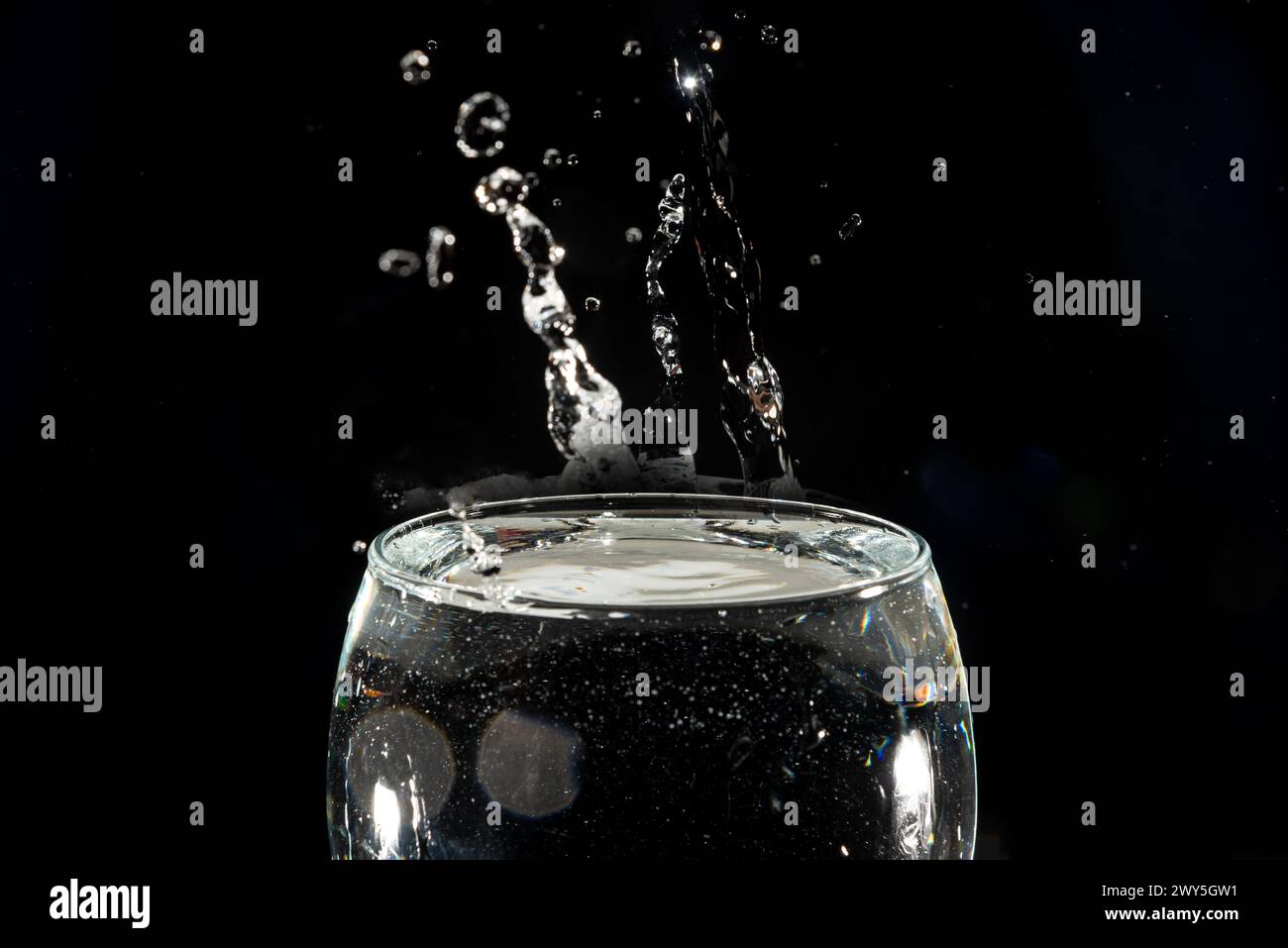 Primo piano di una tazza di vetro trasparente con acqua all'interno e gocce che creano uno spruzzo. Isolato su sfondo scuro. Foto Stock