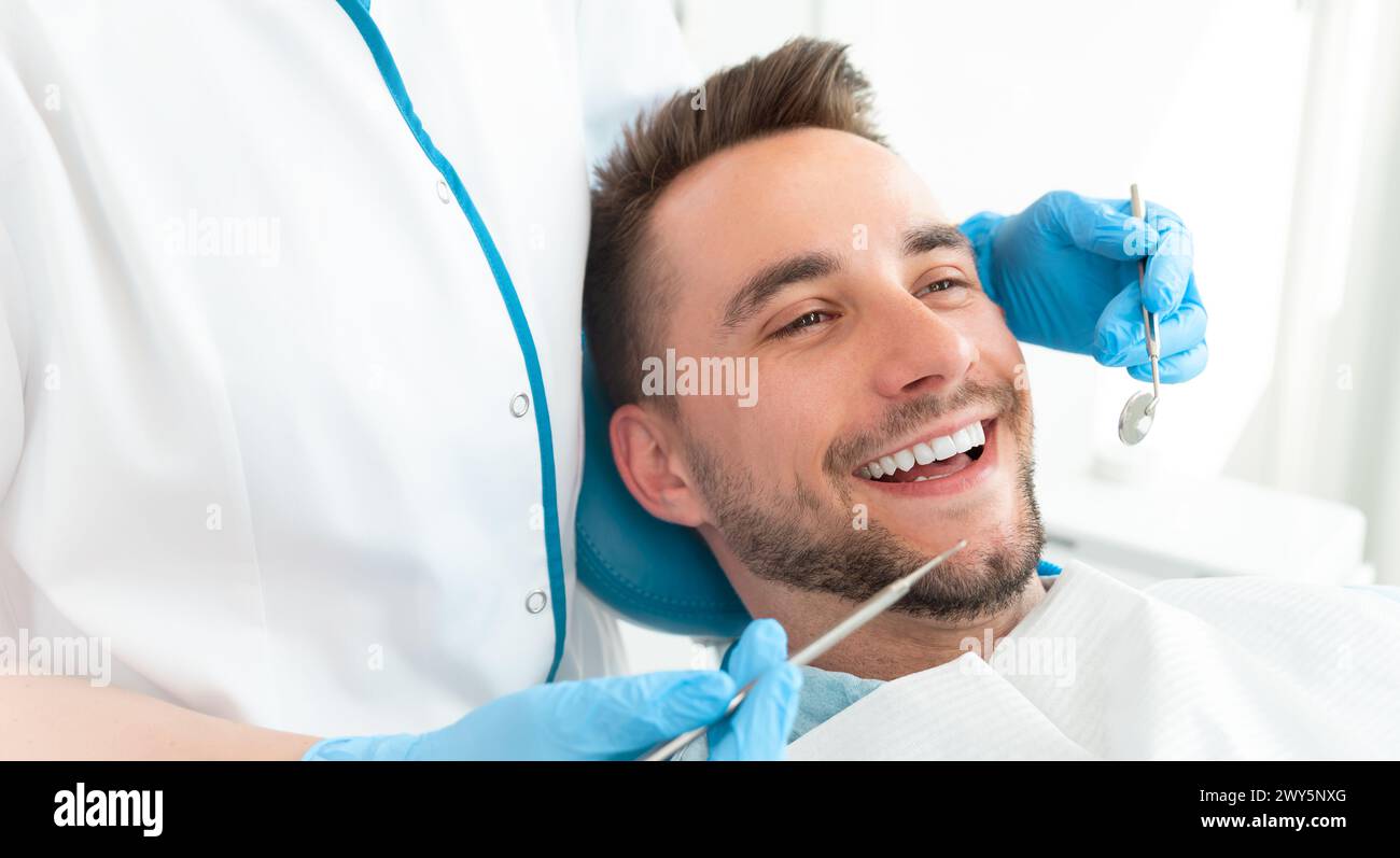 Un uomo sorride mentre un dentista lavora sui suoi denti. Il dentista indossa i guanti blu e tiene un paio di strumenti dentali. Foto Stock
