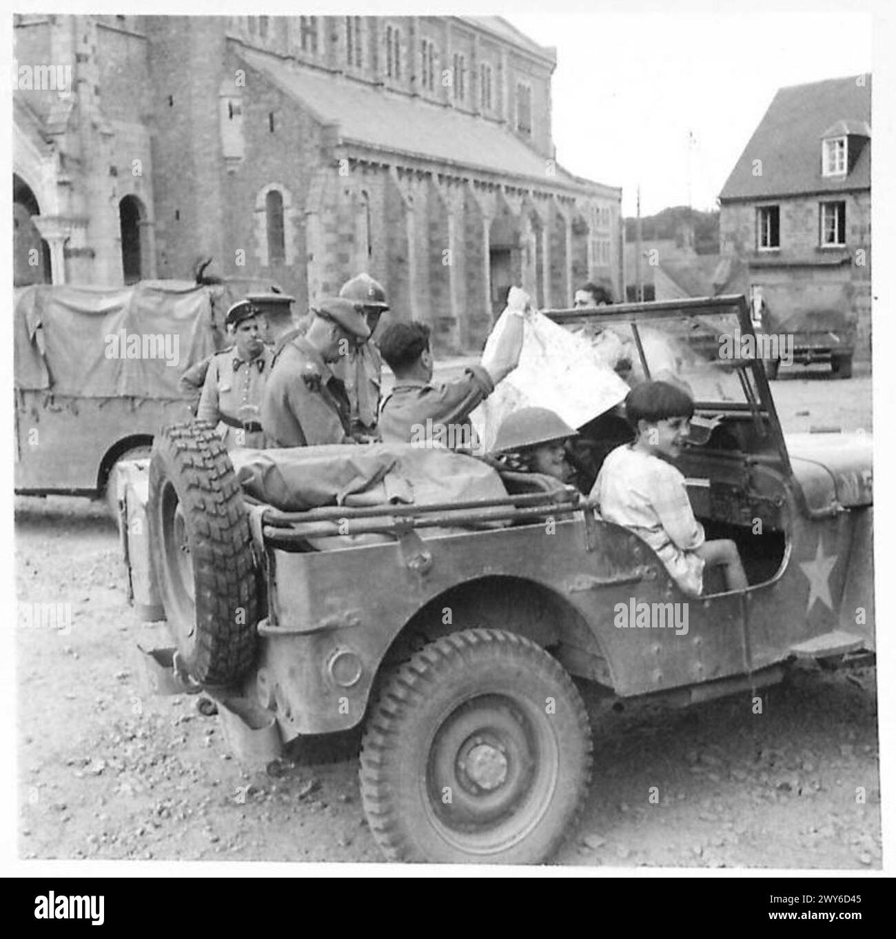 Ufficiali alleati di Gran Bretagna, Francia e Stati Uniti conferiscono in piazza Athis in Normandia mentre i bambini francesi giocano su una jeep. Foto Stock