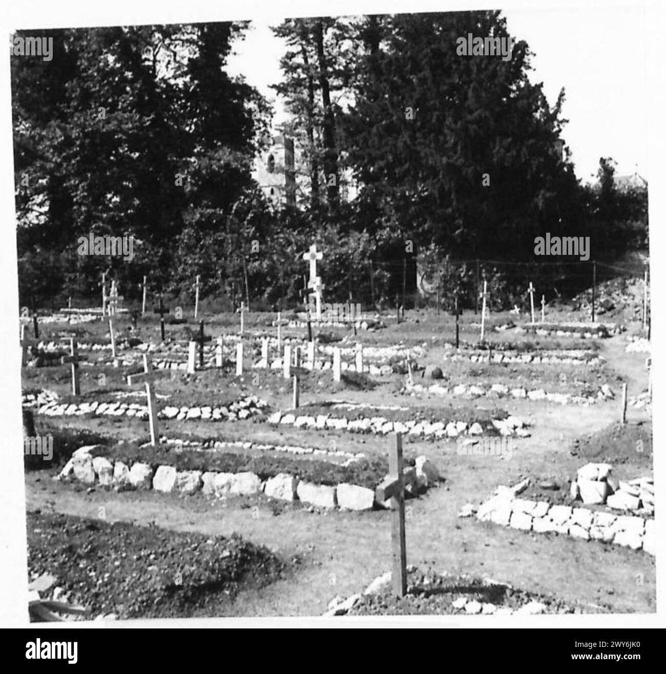 Una sezione completa del cimitero militare di Ranville in Normandia presenta una chiesa e una torre visibili attraverso gli alberi circostanti. Foto Stock