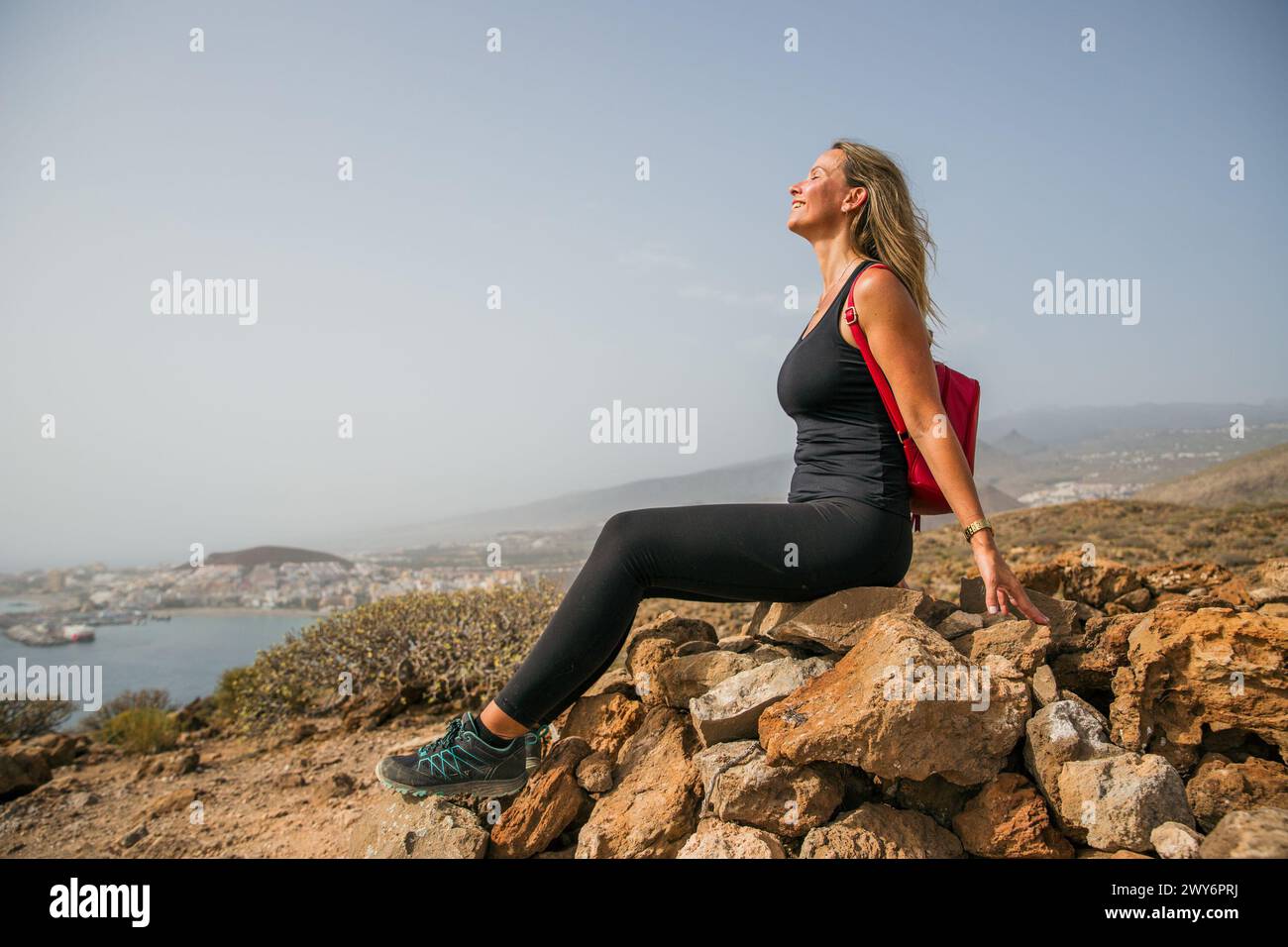 Una donna è seduta su una roccia che si affaccia sull'oceano. Indossa una canotta nera e pantaloni neri Foto Stock
