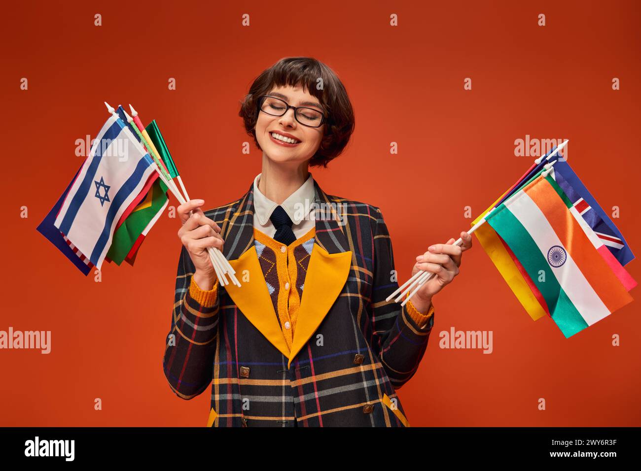 felice ragazza universitaria con l'uniforme e gli occhiali con bandiere multiple e in piedi sullo sfondo arancione Foto Stock