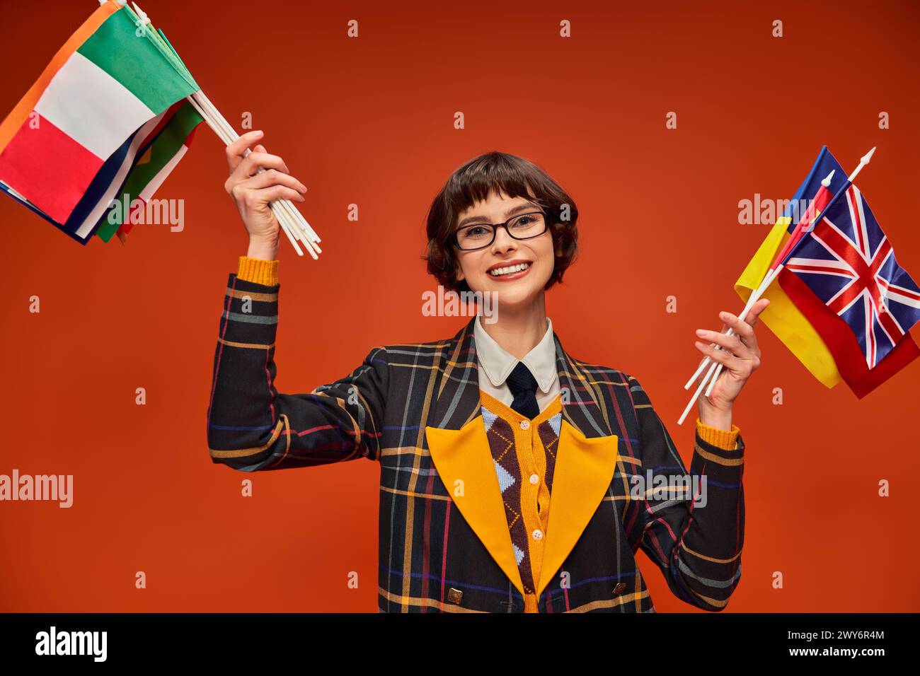 ragazza studentessa felice con l'uniforme e gli occhiali che reggono bandiere multiple e in piedi sullo sfondo arancione Foto Stock