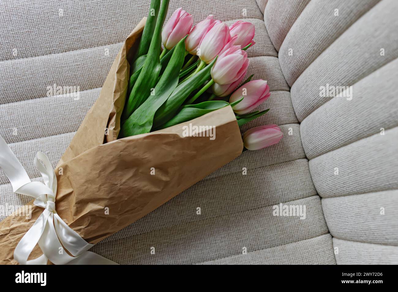Bouquet di tulipani rosa pastello freschi avvolti nella carta artigianale decorata con nastro di raso adagiato sulla sedia beige, Foto Stock