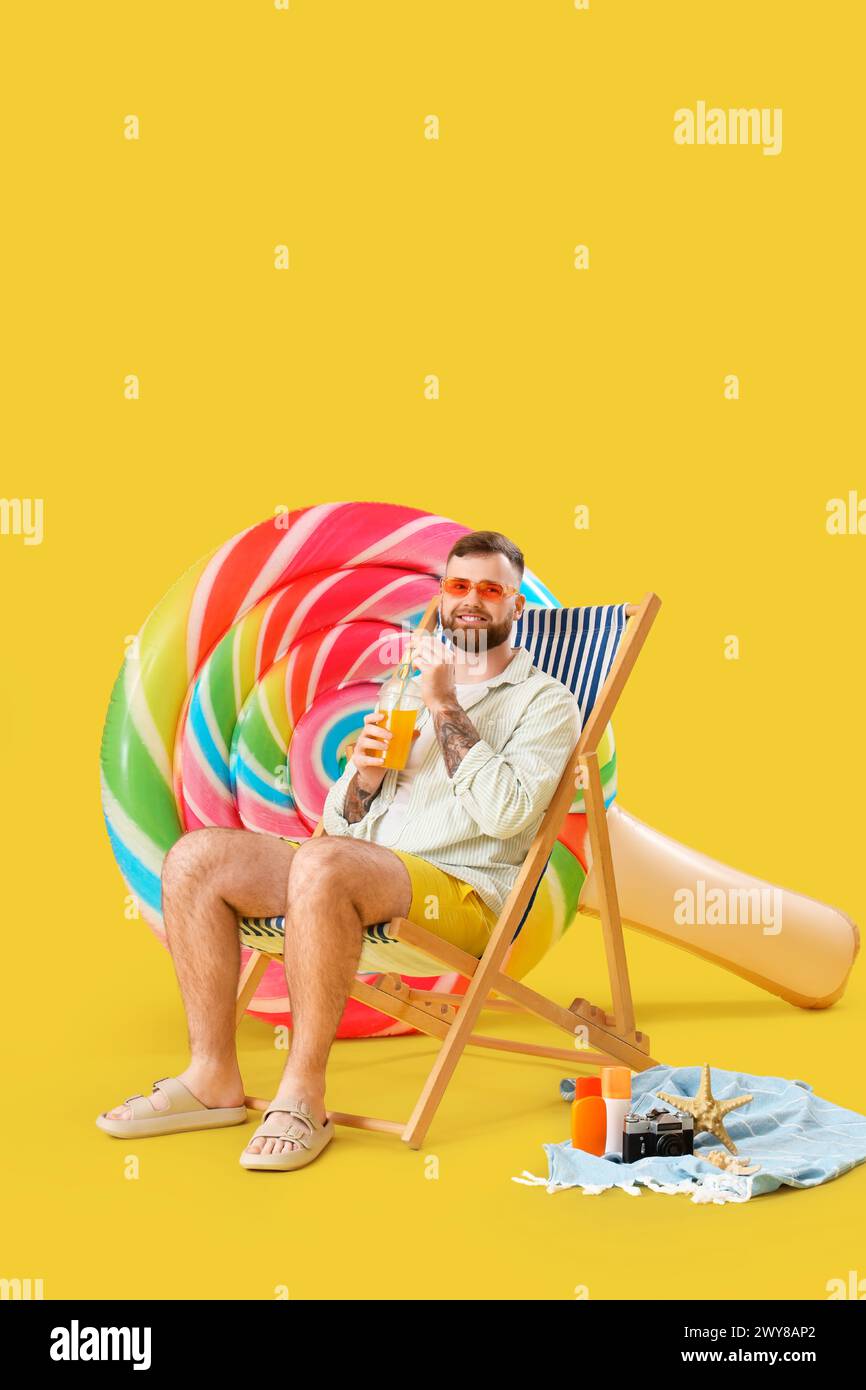 Giovane uomo con materasso gonfiabile a forma di lecca lecca e tazza di succo di plastica in sdraio su sfondo giallo Foto Stock