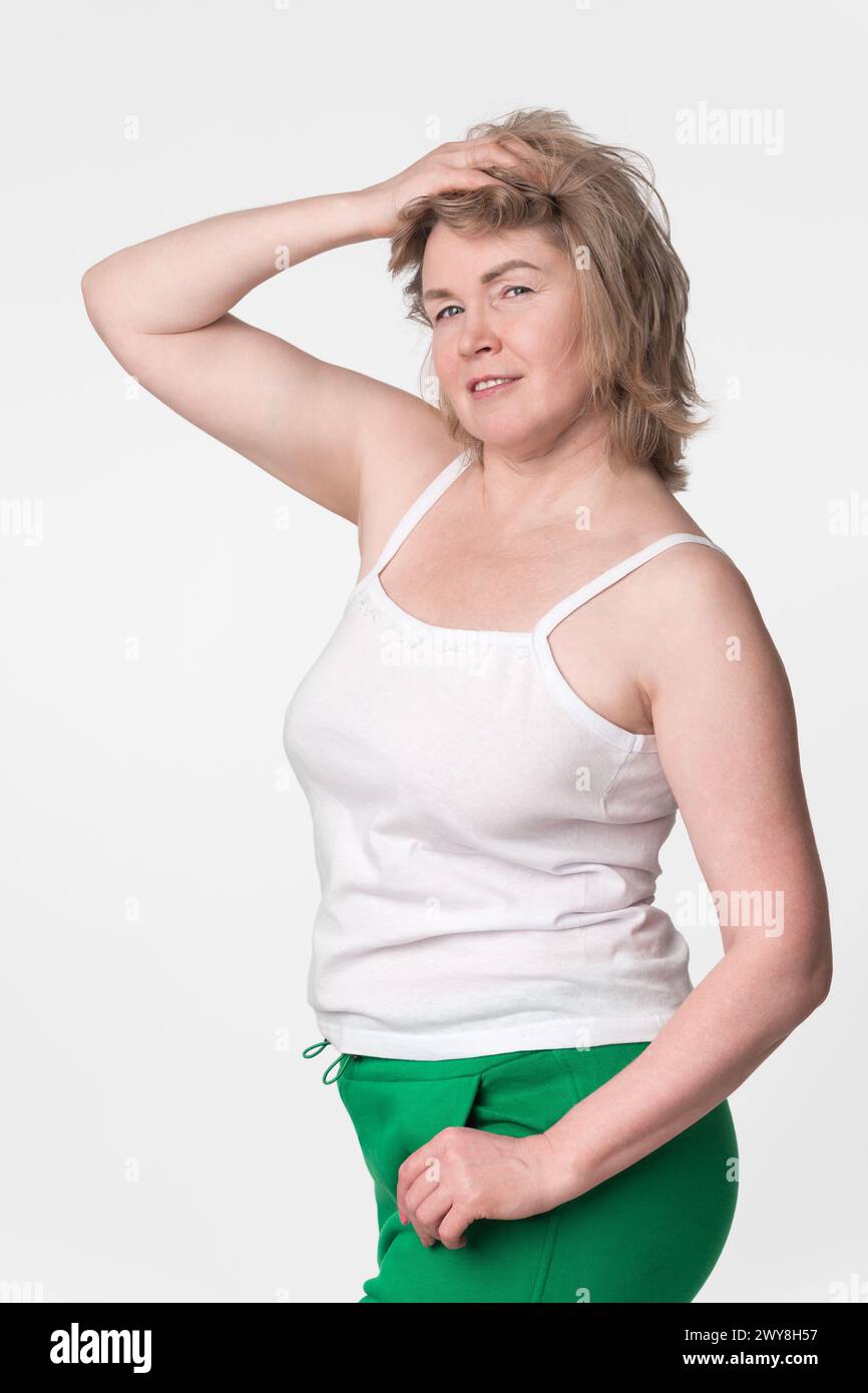 Donna con una figura grintosa in posa con una mano sollevata alla testa e l'altra mano in tasca, in piedi su sfondo bianco. Autentico modello maturo che guarda c Foto Stock