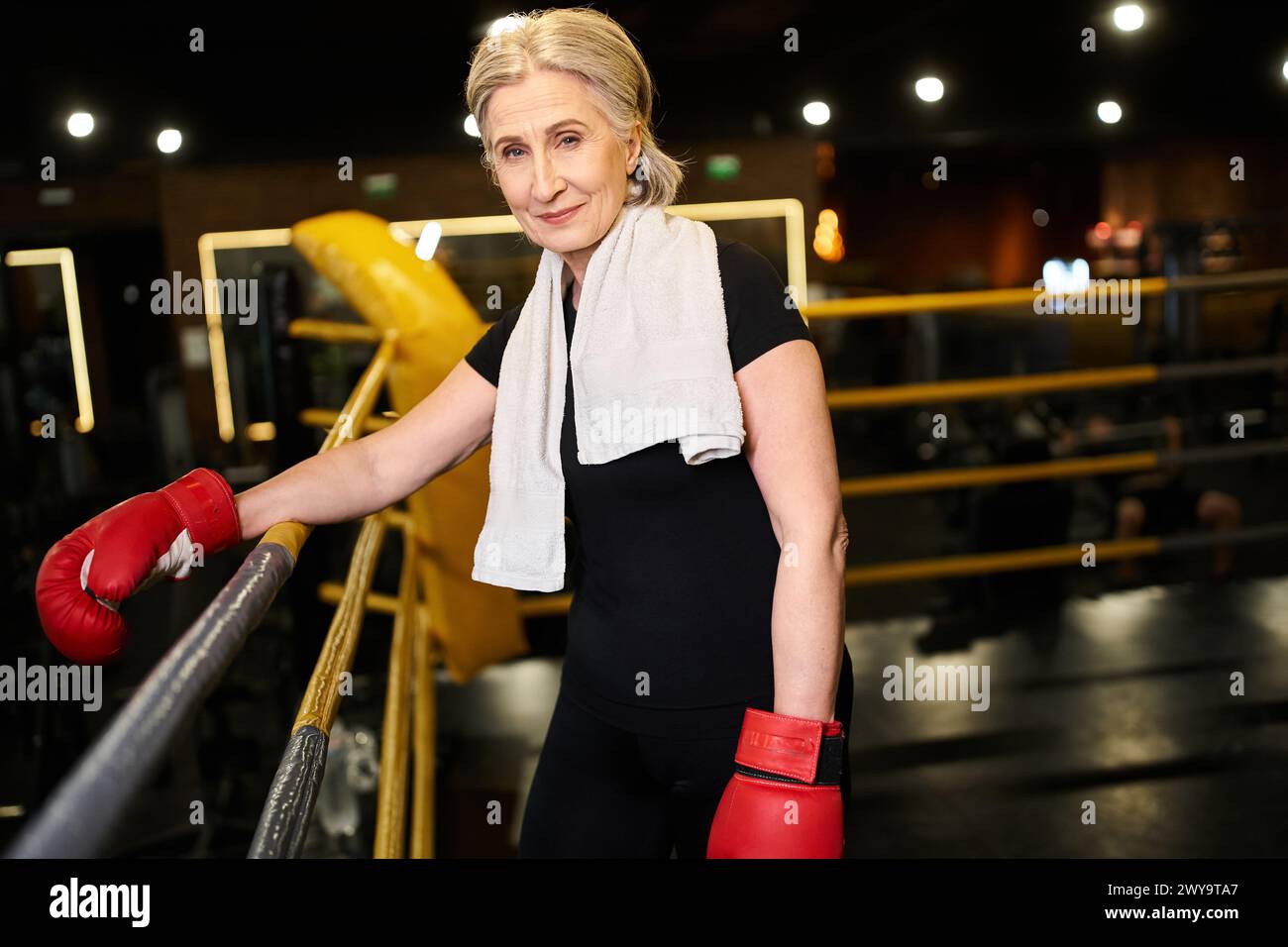 attraente donna sportiva senior gioiosa con asciugamano e guanti da boxe sorridente alla fotocamera sull'anello Foto Stock