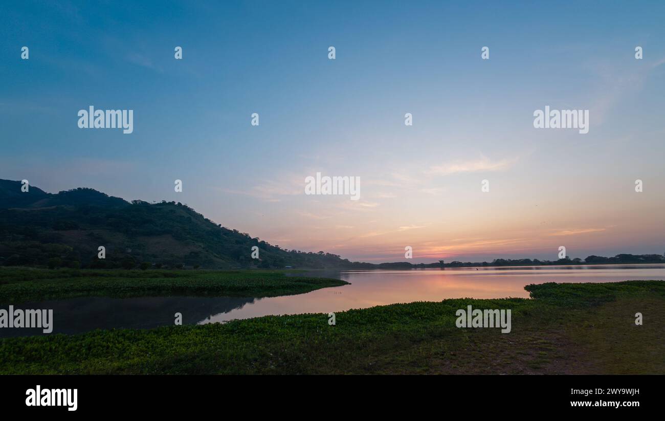 Delicate sfumature pastello di tramonto su un lago tranquillo con colline ondulate e un chiaro riflesso sull'acqua. Foto Stock