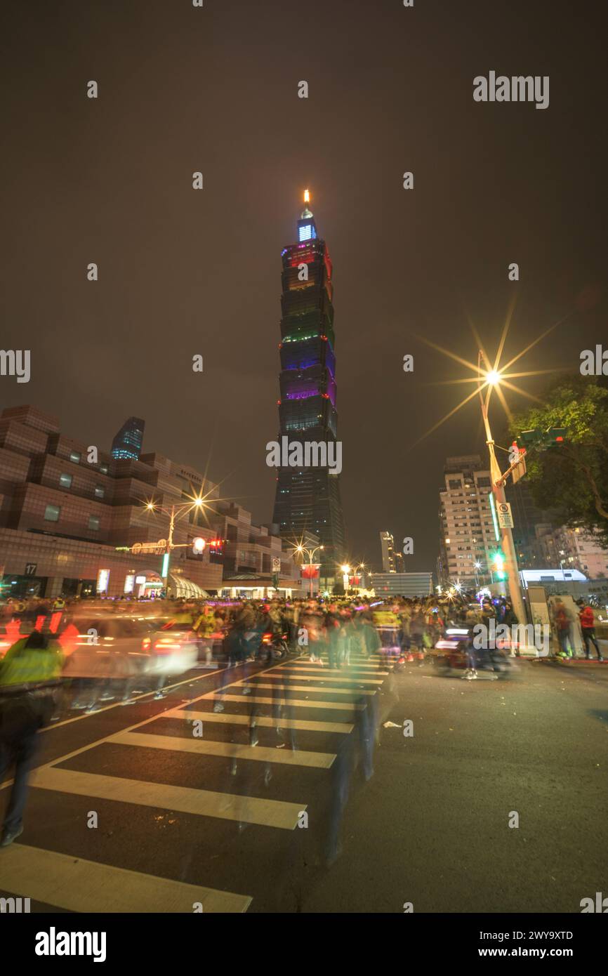 La vivace scena di strada con traffico e pedoni in movimento conduce alla torre illuminata di Taipei 101 contro il cielo scuro dopo lo spettacolo di fuochi d'artificio di Capodanno Foto Stock