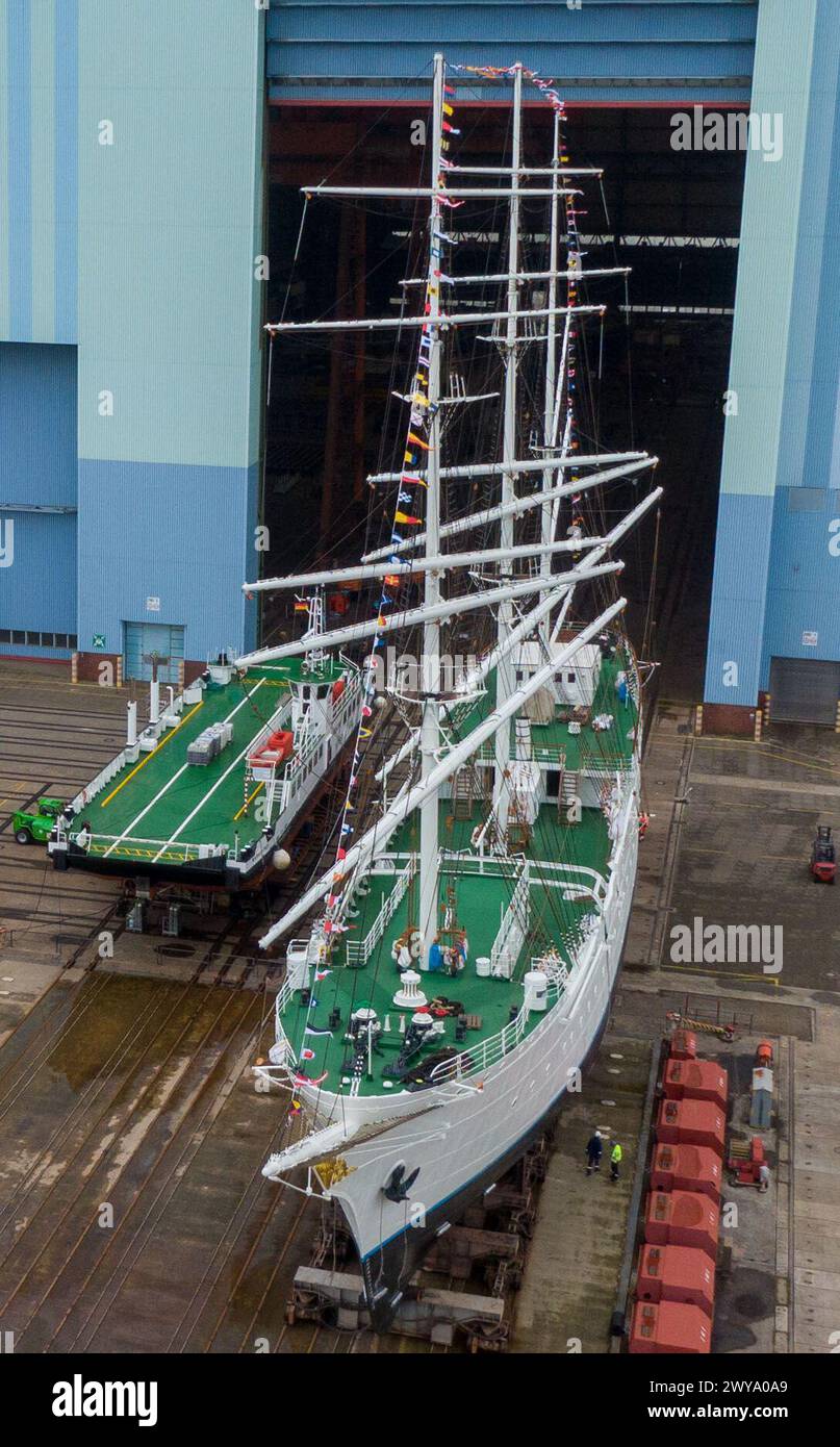 5 aprile 2024, Meclemburgo-Pomerania occidentale, Stralsund: Il "Gorch Fock 1" ha lasciato la sala di costruzione ed è in piedi sull'ascensore della nave (vista aerea con drone). Dopo aver trascorso diversi mesi nella grande sala di costruzione navale del cantiere Volkswerft di Stralsund, il tradizionale veliero "Gorch Fock 1", vecchio di 90 anni, è tornato all'aria aperta. Lo stesso giorno, l'ex nave addestratrice a vela ha lasciato la sala del cantiere con un nuovo, scintillante rivestimento bianco di vernice, dove i costruttori navali hanno effettuato estesi lavori di ristrutturazione. In futuro, la nave sarà nuovamente aperta al pubblico come Foto Stock