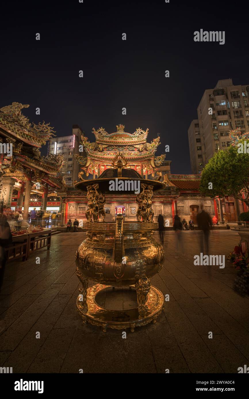 La foto presenta un cancello anteriore del tempio Longshan riccamente decorato con intricate incisioni e animato di fedeli di notte Foto Stock