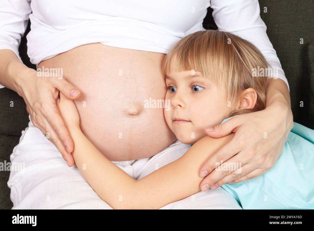 Una ragazza con gli occhi larghi e uno sguardo stupendo abbraccia dolcemente la pancia della madre incinta mentre si siedono insieme su un divano. Momento di legame familiare e un Foto Stock