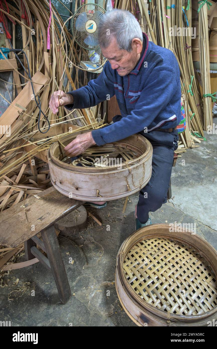 Un anziano artigiano intreccia meticolosamente strisce di bambù per creare un cestino tradizionale nella sua bottega nella città di Lukang Foto Stock