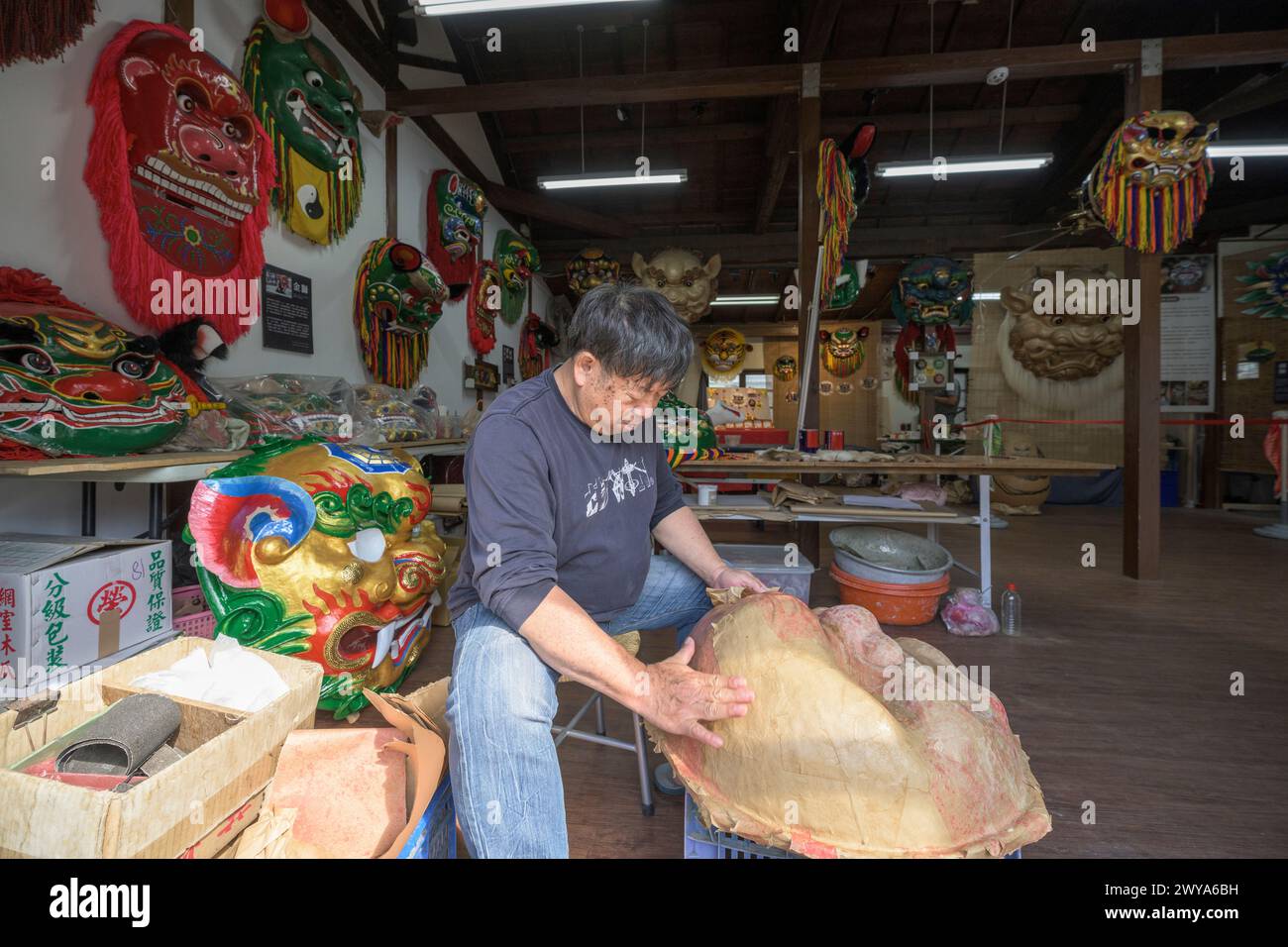 Un artigiano sta realizzando a mano una maschera cinese in un laboratorio illuminato dal sole circondato da maschere e decorazioni colorate Foto Stock