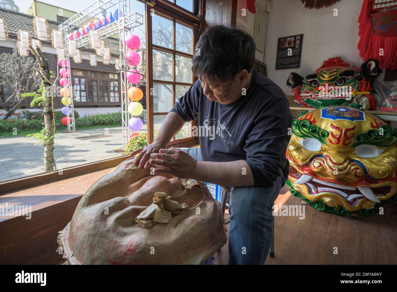 Un artigiano sta realizzando a mano una maschera cinese in un laboratorio illuminato dal sole circondato da maschere e decorazioni colorate Foto Stock