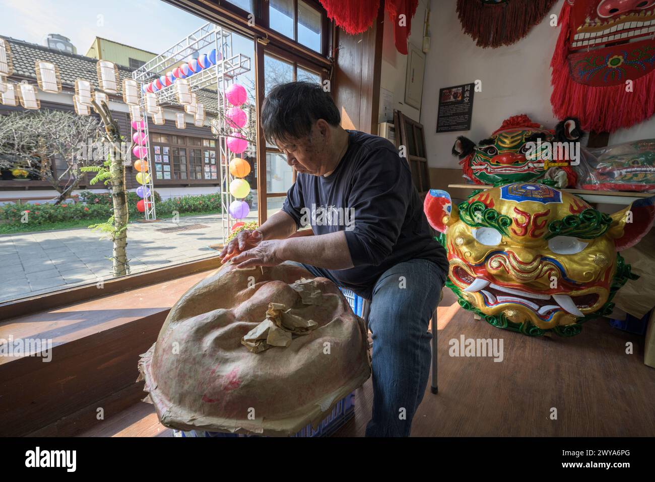 Un artigiano sta realizzando a mano una maschera cinese in un laboratorio illuminato dal sole circondato da maschere e decorazioni colorate Foto Stock