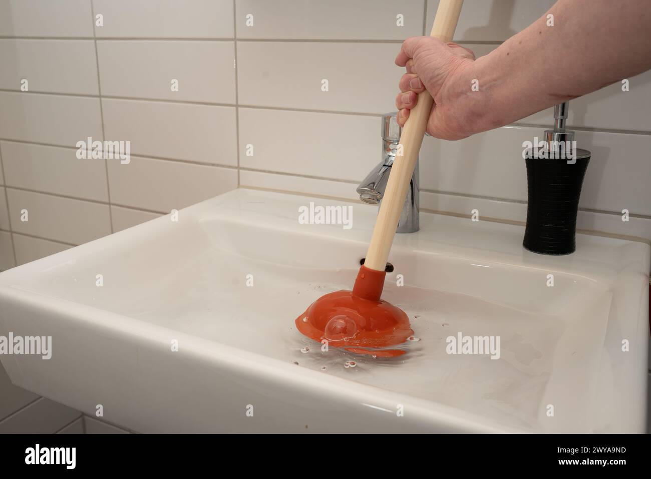 Uomo che utilizza lo stantuffo per rimuovere l'ostruzione del tubo di scarico sul lavandino del bagno. Si vedono solo la mano e il braccio dell'uomo. L'acqua nel lavandino mostra agitazione. . Foto Stock