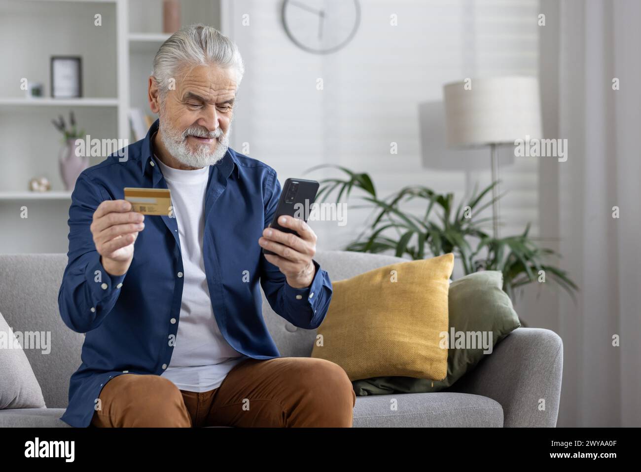 Uomo con barba matura che utilizza comodamente lo smartphone e la carta di credito per fare acquisti online da casa. Foto Stock