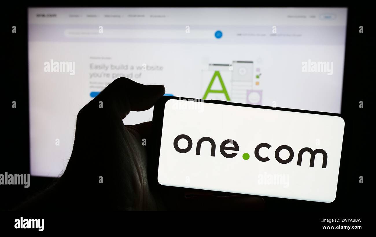 Persona che detiene un telefono cellulare con il logo della società di web hosting One.com Group davanti alla pagina web aziendale. Mettere a fuoco il display del telefono. Foto Stock