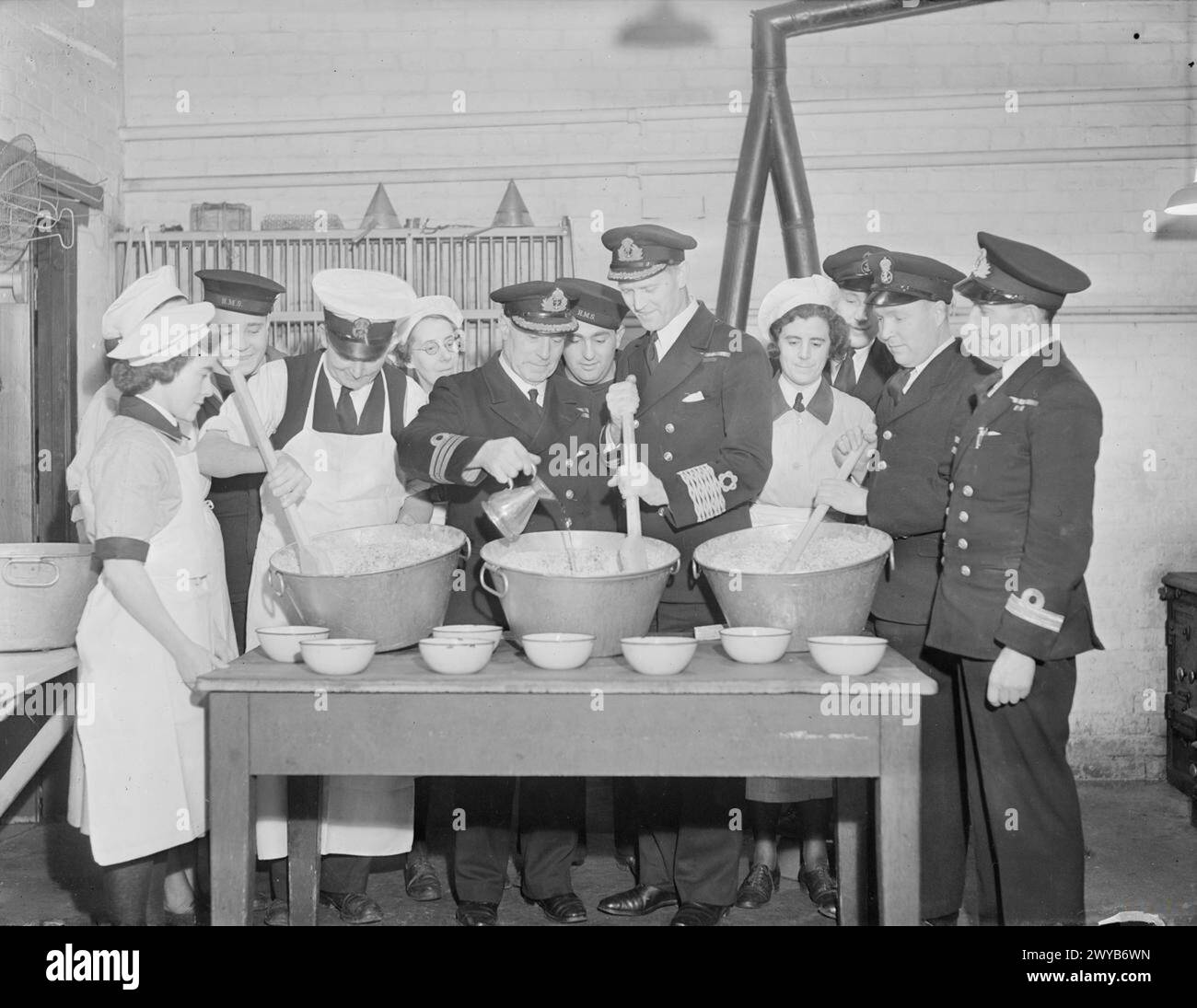 BUDINO DI NATALE DELLA MARINA IN PREPARAZIONE. 1 DICEMBRE 1943, HARWICH. LA TRADIZIONALE MESCOLANZA DEL BUDINO DI NATALE. RATING E WRENS HANNO PRESO PARTE E L'EMOZIONE FINALE È STATA DATA DAL CONTABILE DI BASE. - Il comandante di base, CDR W H Beedle, RN, che versa nel rum mentre l'ufficiale contabile di base paga il capitano e D Guiness, RNR mescola il budino. , Foto Stock