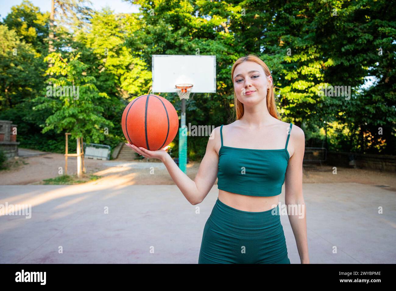 Una donna sta tenendo un basket in un parco. Il parco è circondato da alberi e dispone di un campo da pallacanestro. La donna sorride e le piace l'hersel Foto Stock
