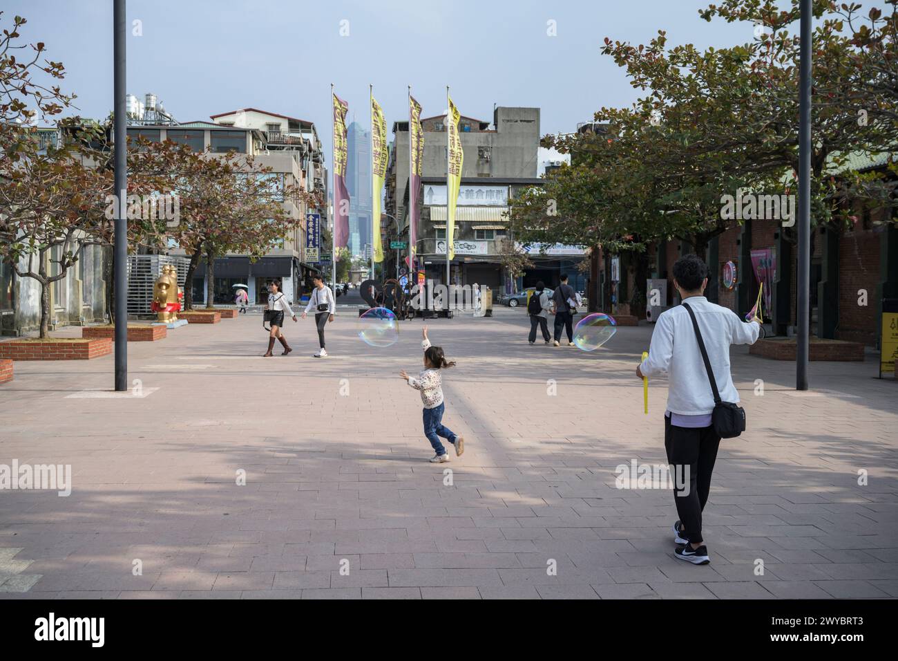 Un bambino corre dietro le bolle di sapone in una spaziosa piazza urbana circondata da persone ed edifici cittadini Foto Stock