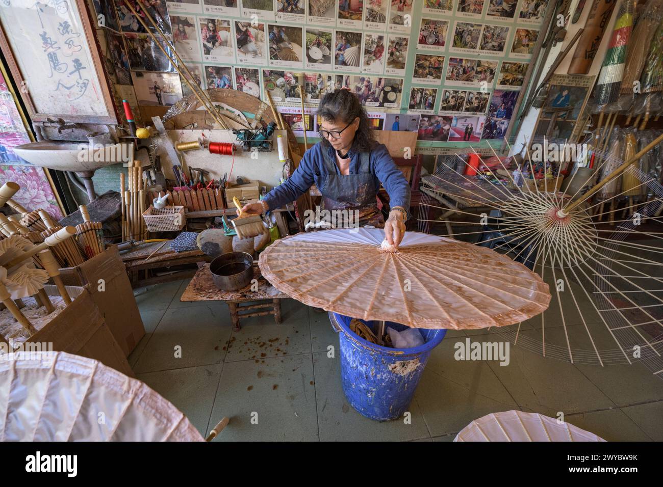 In un laboratorio, un artigiano si concentra sulla creazione di ombrelli cinesi tradizionali circondati da prodotti finiti Foto Stock