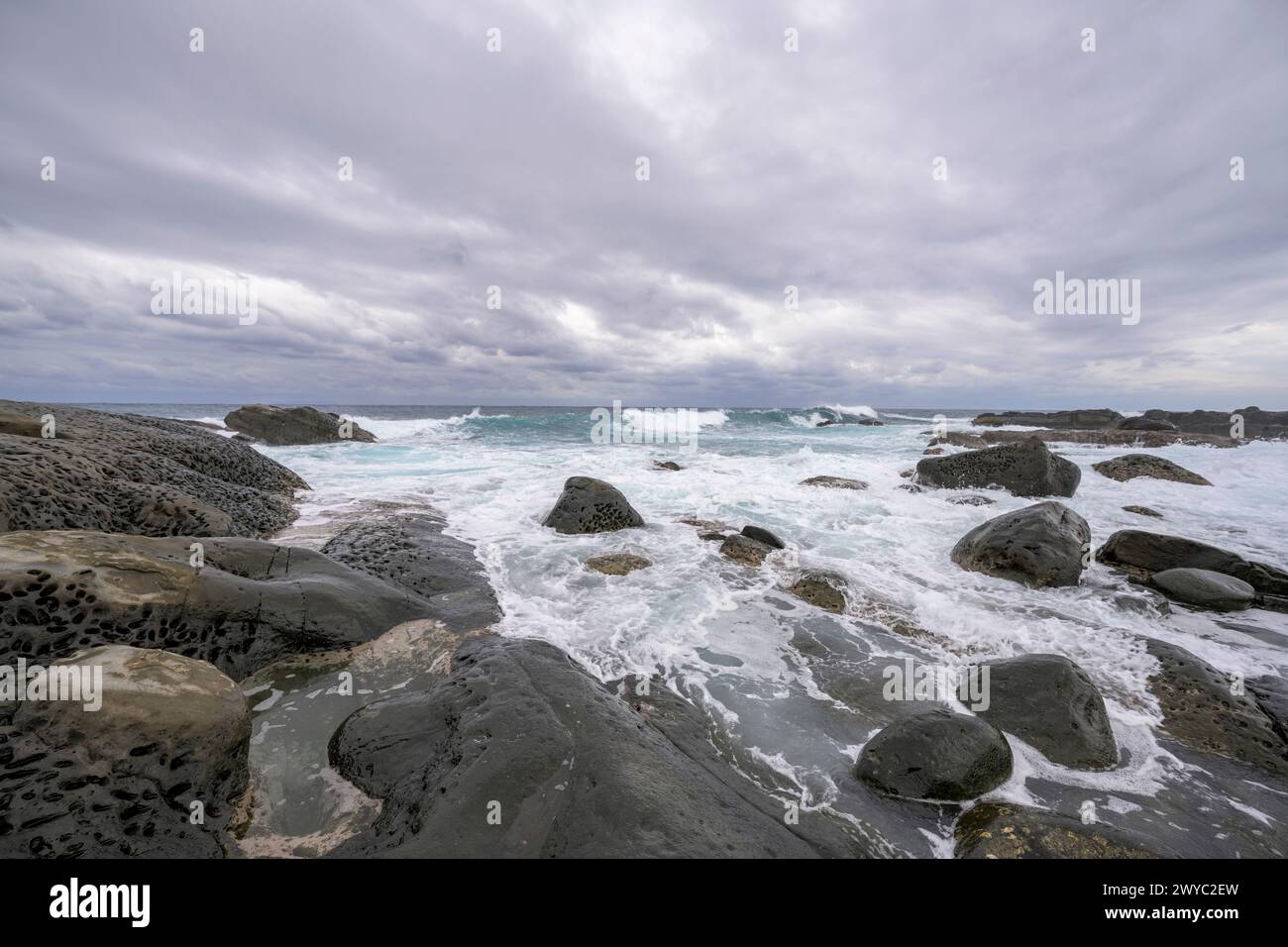 Una giornata ricoperta su una costa rocciosa che mostra schemi unici di fori erosivi con onde aspre sullo sfondo Foto Stock