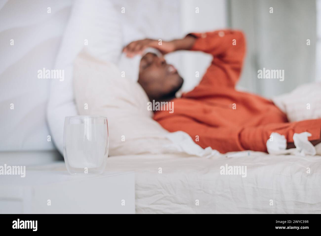 Uomo malvagio sdraiato a letto con acqua e tessuti, concetto di malattia a casa. Un uomo afroamericano giace a letto con il braccio sulla fronte, un bicchiere d'acqua in primo piano, catturando un momento di riposo durante la malattia Foto Stock