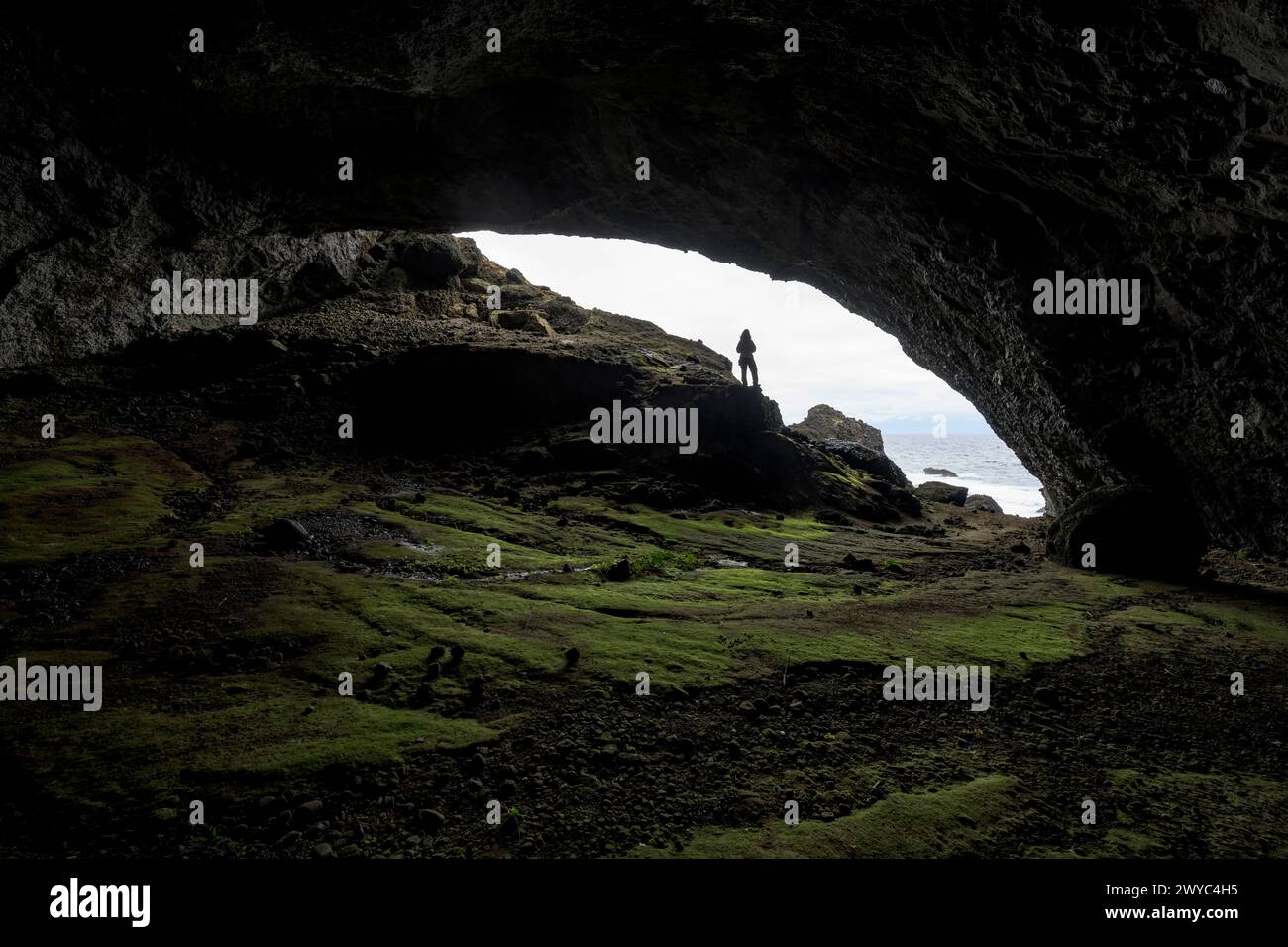 Una persona si trova alla foce di una grande grotta marina, sagomata contro il cielo luminoso e la costa frastagliata Foto Stock
