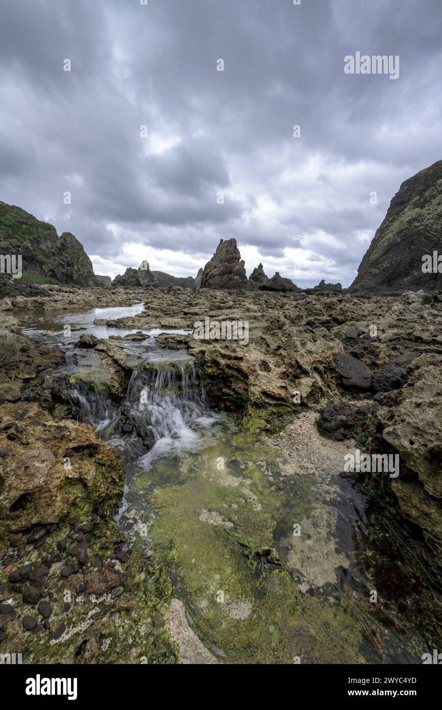 Una dolce cascata scende da rocce rocciose in piscine con maree con uno sfondo di aspre formazioni rocciose costiere Foto Stock