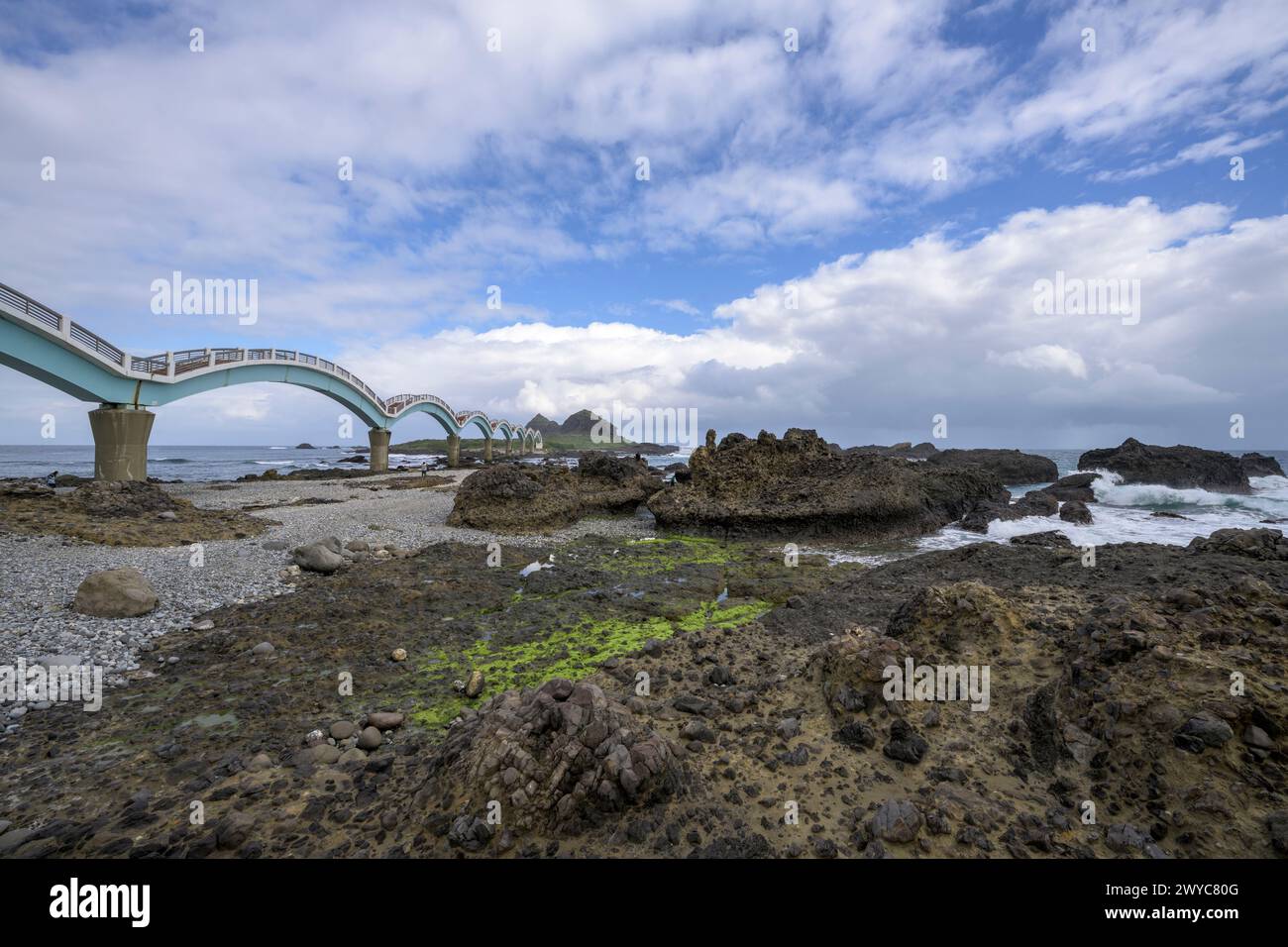 Uno splendido ponte dell'Arco di Sanxiantai si estende su una scena costiera rocciosa con onde e un cielo blu stratificato di nuvole Foto Stock