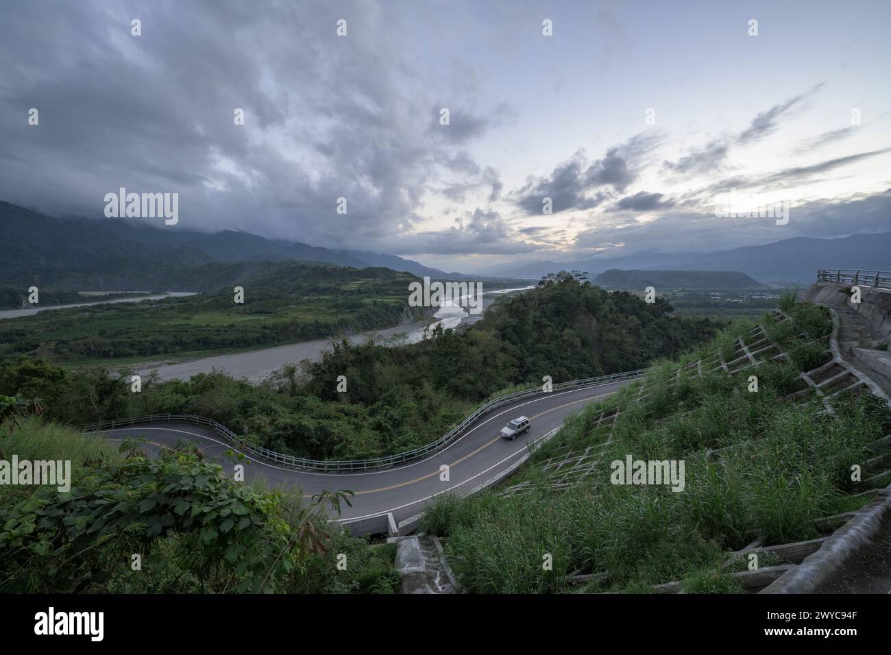 Un'immagine suggestiva che mostra la curva naturale di un fiume con un'adiacente strada tortuosa che taglia maestose montagne contro un cielo nuvoloso al crepuscolo Foto Stock