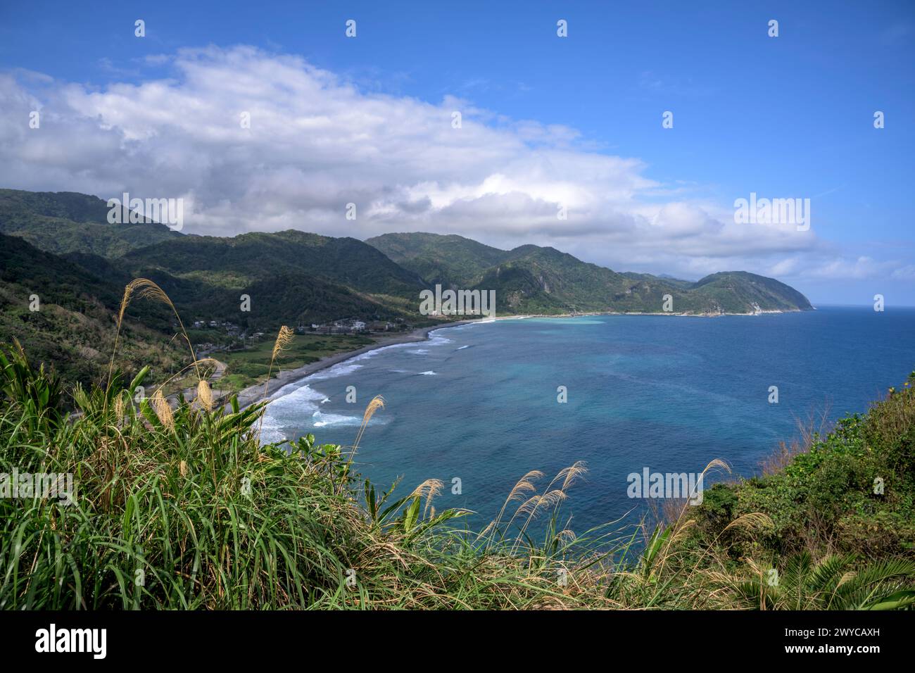 Un ampio paesaggio costiero con un piccolo villaggio sul mare accoccolato tra lussureggianti colline sotto un cielo luminoso Foto Stock